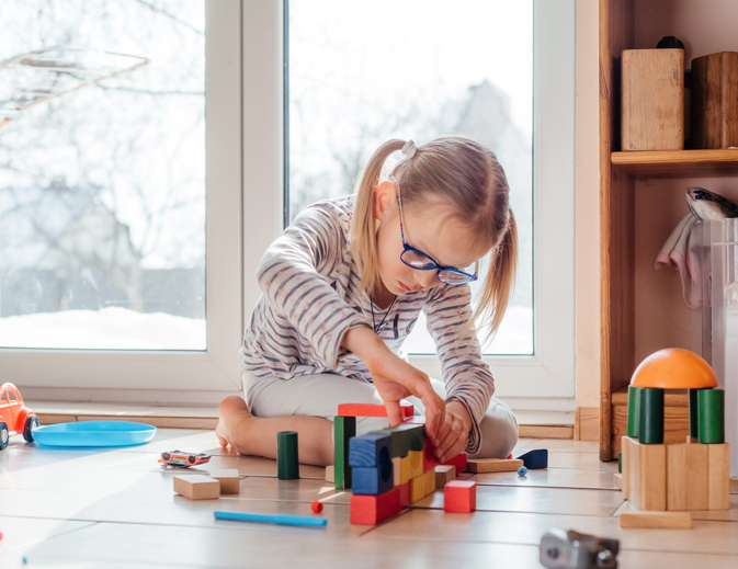 A little girl in eyeglasses playing with wooden building blocks at home