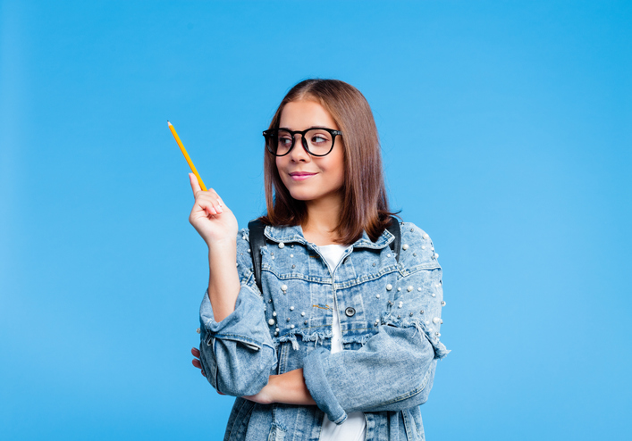Happy teenager wearing oversized denim jacket, white t-shirt and nerdy black eyeglasses pointing with pencil at copy space. Smiling female high school student standing against blue background.