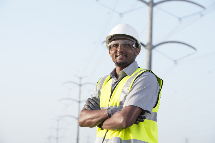 Portrait of manual worker in Personal protective equipment (PPE) standing under high voltage transmission lines against tubular transmission towers.