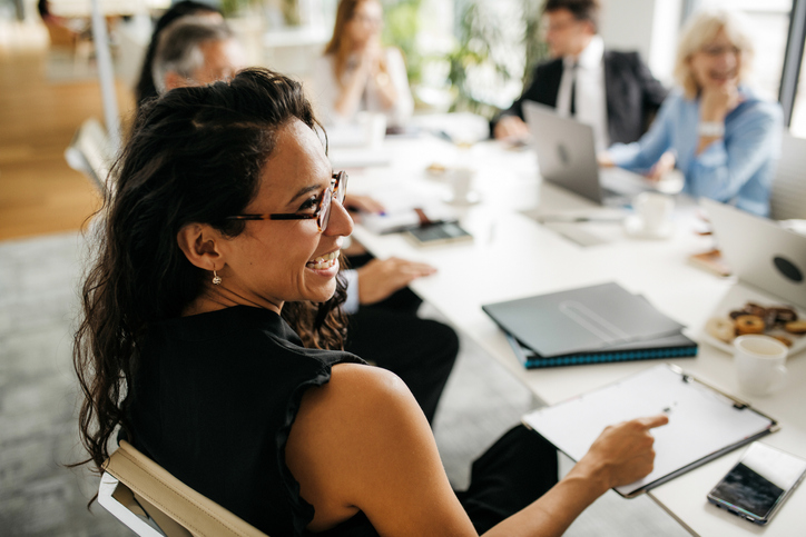 Over the shoulder profile of bespectacled female executive in early 30s sitting at conference table and laughing as she interacts with off-camera colleague.