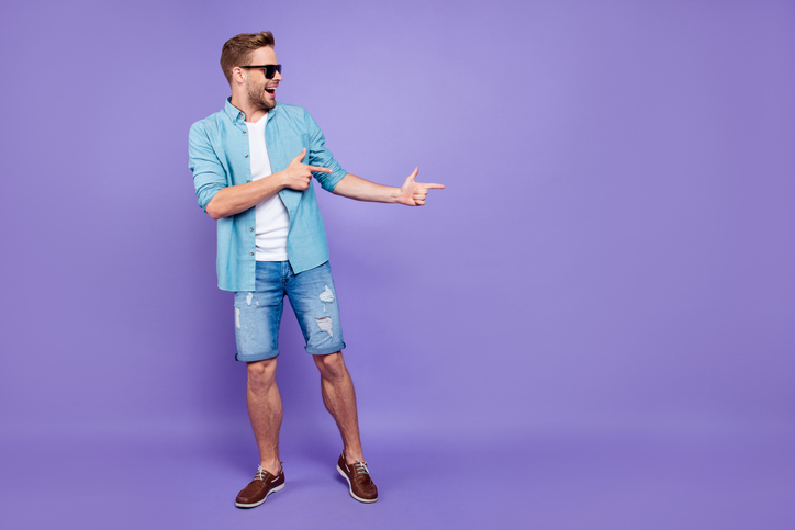Full body size length of attractive bearded trendy stylish confident cheerful man, wearing denim and glasses, pointing with fingers aside, over bright violet purple background