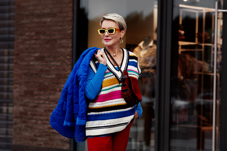 Fashion Street style autumn or spring. Model is wearing blue jacket and sweater, striped gilet, red pants and yellow sunglasses. Woman posing outside clothing store