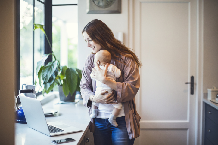 Smiling mother using laptop while carrying baby at kitchen counter. Woman is working while holding son at home.