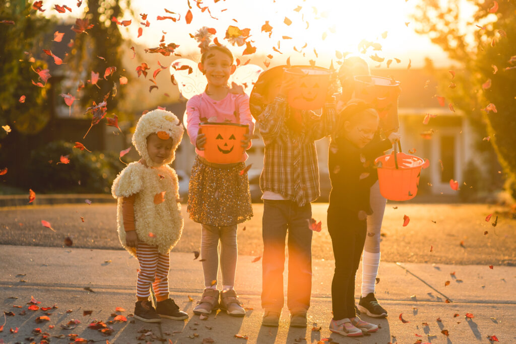 Five children of varying ages, ethnicities, and costumes go trick or treating. They include a cowboy, a duck, a fitness instructor, a gymnast, and a black cat. They are holding candy buckets in a residential neighborhood and they're throwing leaves in the air.