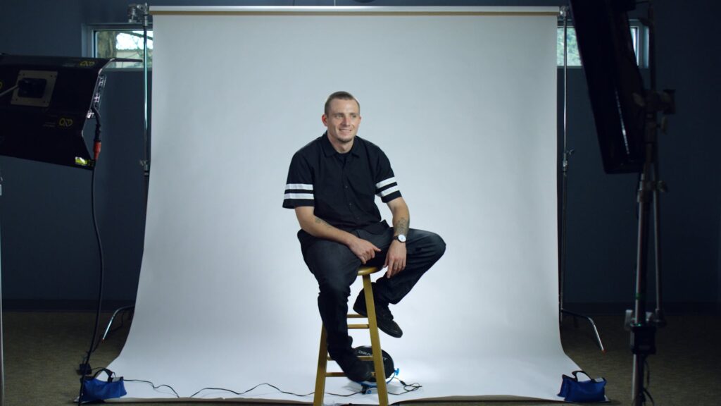 A man in front of a while backdrop sitting on a stool